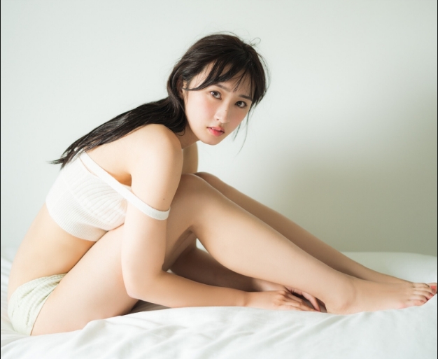 A young woman in a light-colored outfit sitting on a bed with a soft expression, showcasing a natural and fresh look.