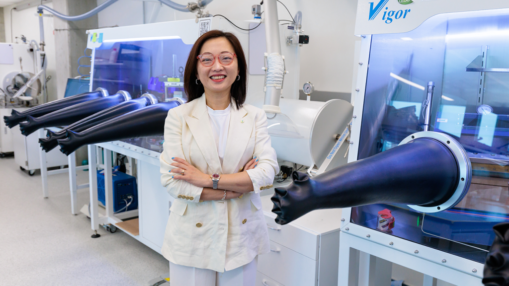 研究人員在實驗室中,穿著白色西裝,微笑著站在設備旁。背景顯示多個儀器和黑色手套箱。