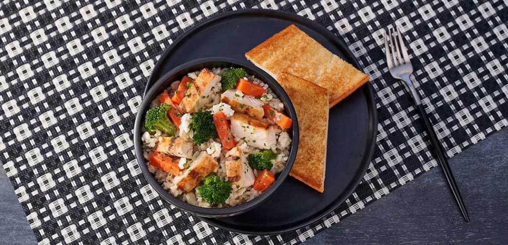 一碗健康的炒飯，包含烤雞肉、青花菜、胡蘿蔔等新鮮蔬菜，旁邊放著幾片烤吐司，背景是黑白圖案的桌布。