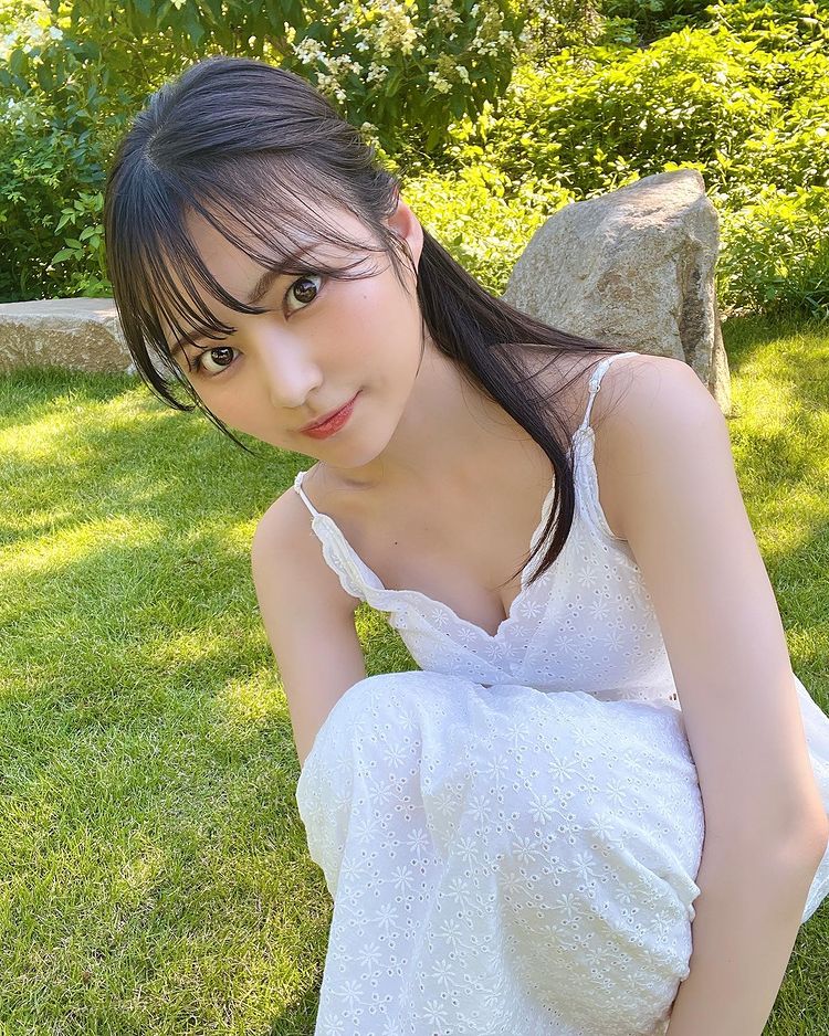 A young woman with long black hair styled in soft waves, wearing a white dress, smiles while sitting on green grass in a garden setting.
