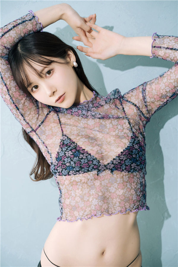 A model posing with arms raised, wearing a sheer floral top that reveals a black bralette beneath, set against a light blue background.