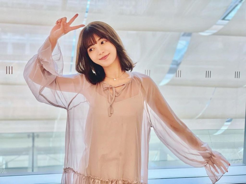 A young woman poses playfully with a peace sign, wearing a flowy, light-colored dress, against a backdrop of soft, natural light.