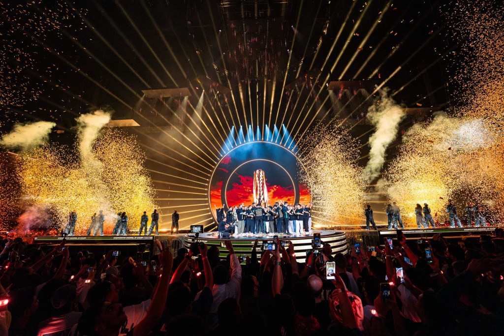 一場盛大的電競比賽開幕儀式，舞台上出現壯觀的燈光和煙火，觀眾群激動拍照。