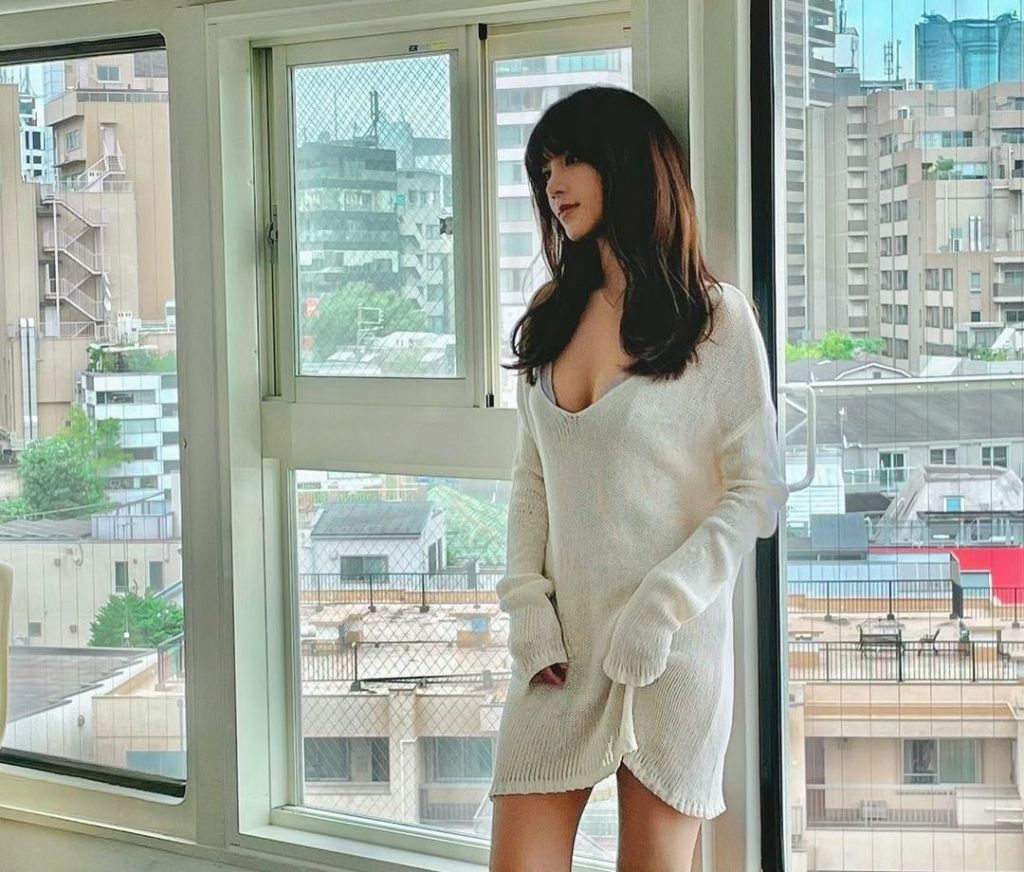 A woman with long dark hair wearing a white oversized sweater stands by a window, looking thoughtfully outside at the cityscape.