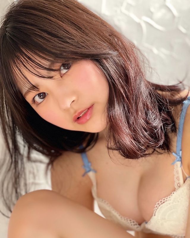 A close-up portrait of a young woman with long, dark hair and natural makeup, wearing a light blue bra with lace details, looking directly at the camera with a gentle expression.