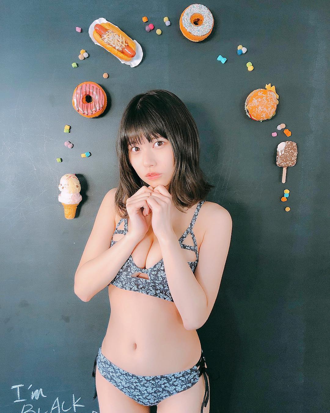 A young woman poses in a floral bikini, standing in front of a black wall decorated with colorful donut and candy props.