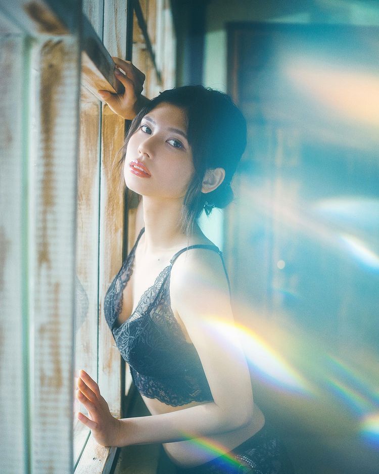 A woman in a lace black bralette poses thoughtfully by a window, with soft light and reflections creating a dreamy atmosphere.
