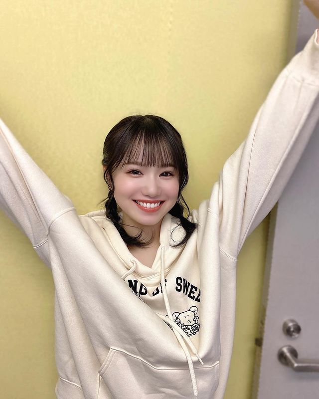 A young woman smiling widely with her arms raised, wearing a beige hoodie with a graphic design, standing against a yellow wall.