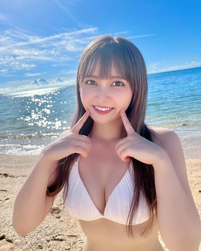 在海灘上微笑的年輕女性，穿著白色泳衣，手指著自己的臉頰，背景是湛藍的海水和晴朗的天空。