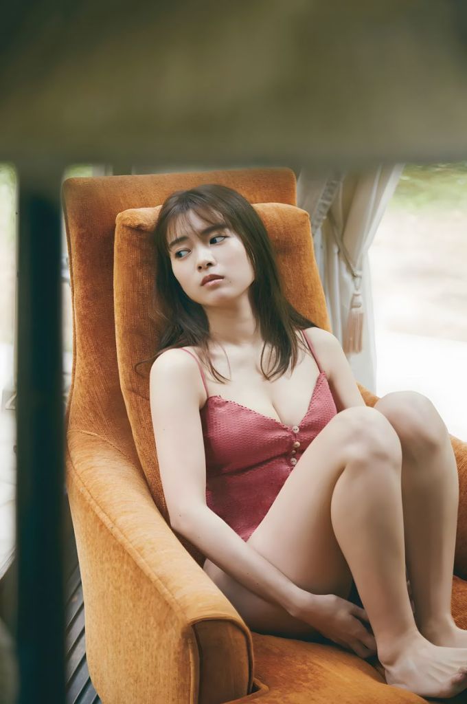 A young woman sitting in an orange armchair, looking pensive while wearing a red lingerie outfit.
