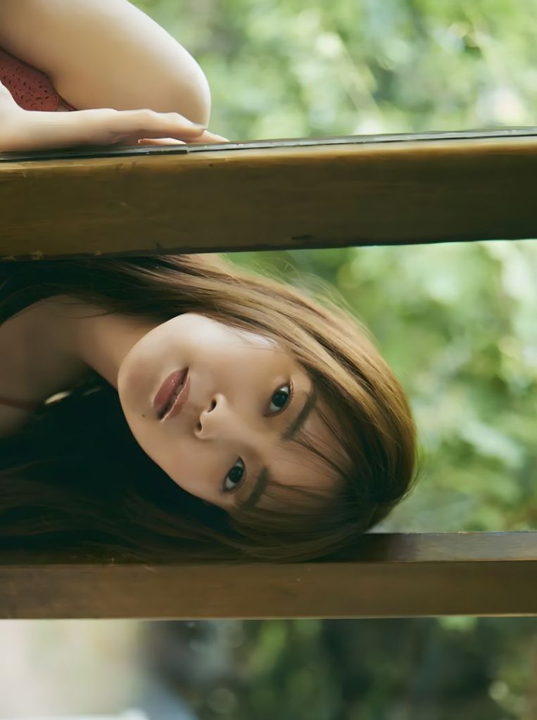 A woman with long hair is resting her head upside down on a wooden surface, surrounded by greenery.