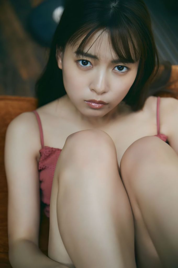 A young woman sitting with her knees drawn up, wearing a pink top, looking directly at the camera with a thoughtful expression.