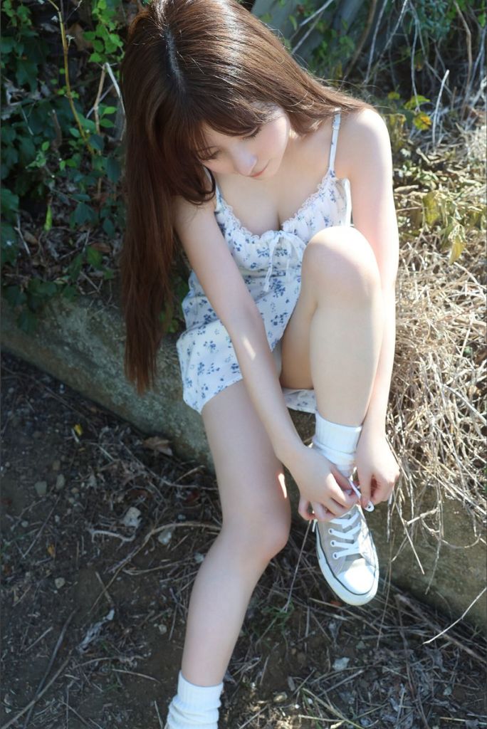 A young person sitting on a rock in a natural setting, tying shoelaces while wearing a light floral dress and ankle socks.