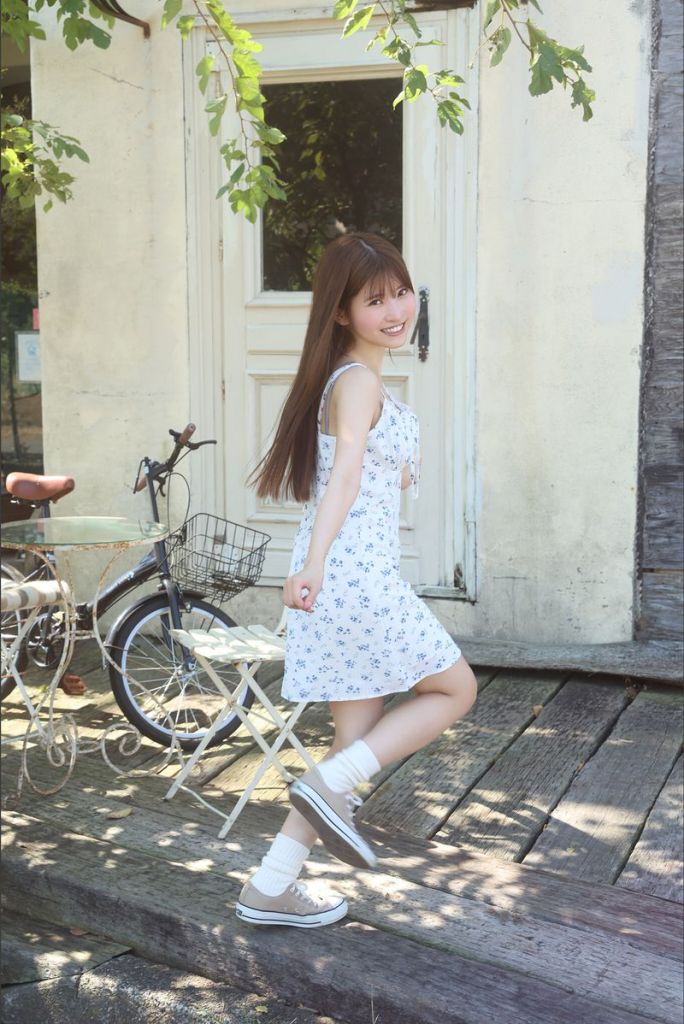 A young woman wearing a floral dress and sneakers is happily posing outdoors, standing on wooden planks with a vintage bicycle nearby and a door in the background.