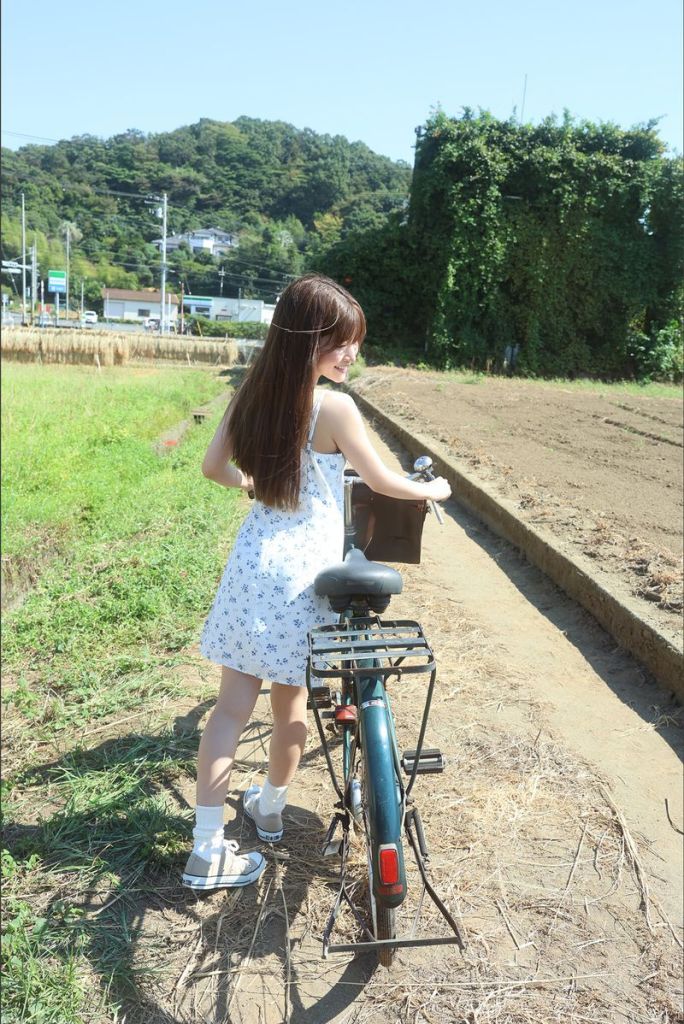 A young woman in a floral dress stands next to a bicycle in a field, with green hills and trees in the background.