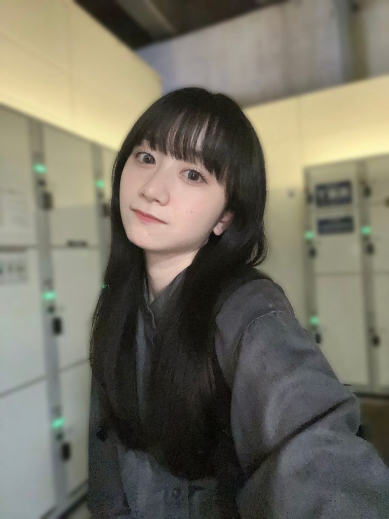 一名年輕女性在閉鎖式空間中自拍，背景有一些儲物櫃，女性擁有長黑色頭髮，穿著深色服裝，面帶微笑。