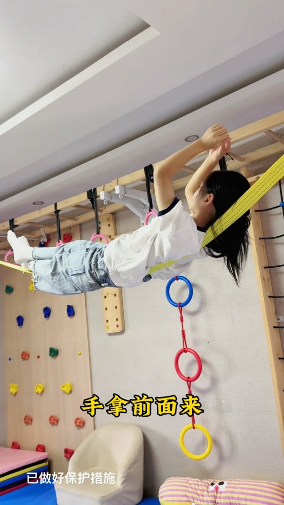 一名九歲女孩在繩索上穩定地躺著，手抓住繩索，展現出驚人的平衡能力，背景為色彩繽紛的健身房設施
