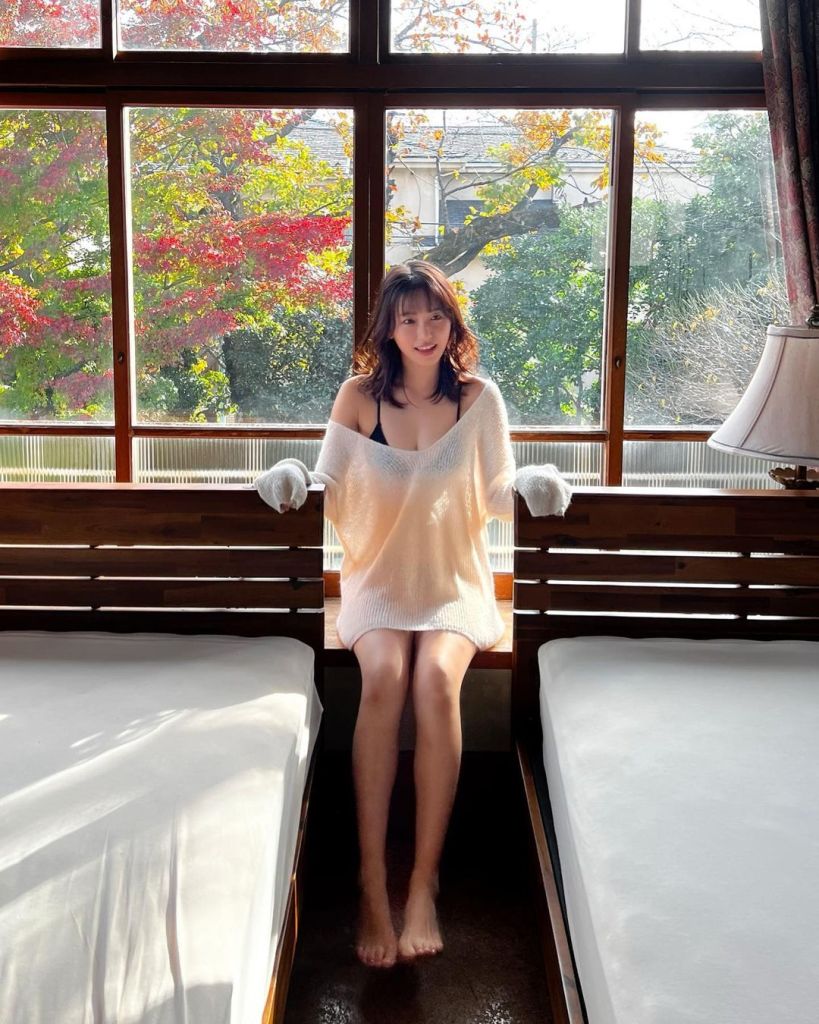 A young woman sitting between two beds in a cozy room, wearing an oversized white sweater and smiling at the camera, with large windows showing a colorful autumn scenery outside.