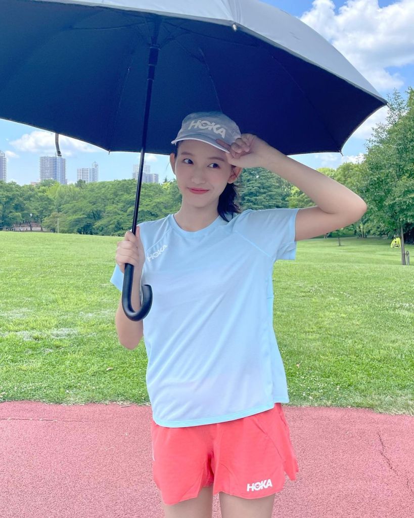 A young woman holding an umbrella while wearing a light blue athletic shirt and pink shorts, standing on a grassy field with a red track in the foreground and city buildings visible in the background.
