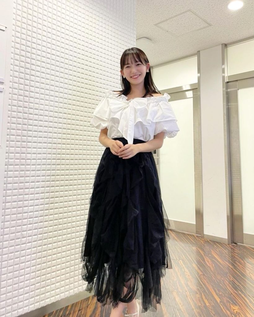 A person is smiling while wearing a white off-the-shoulder top and a black layered skirt, standing in a well-lit interior space with a textured white wall.