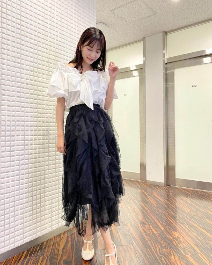 A person in a stylish outfit consisting of an off-the-shoulder white top and a black ruffled skirt, poses gracefully in a modern indoor setting.