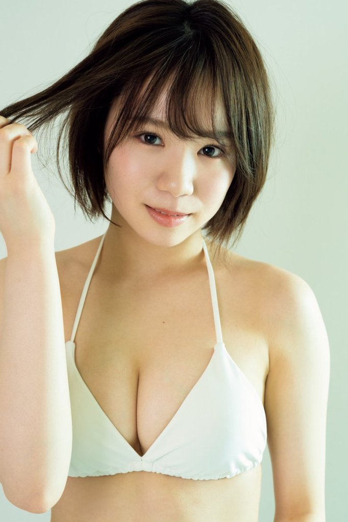 A young woman with short hair is smiling and holding a strand of her hair while wearing a white bikini top against a light background.