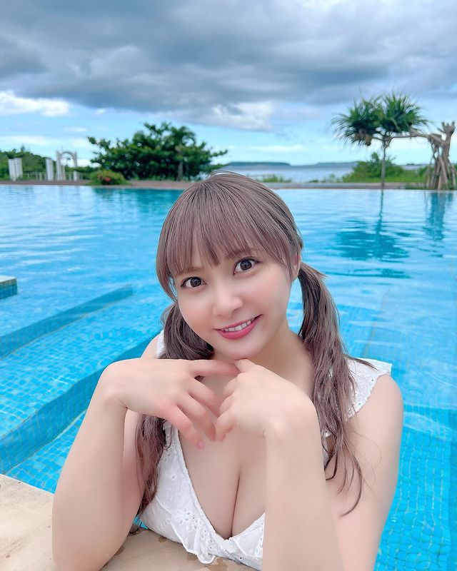 一位穿著白色泳裝的女性在泳池邊微笑，背景是一片美麗的藍天和翠綠的植物。