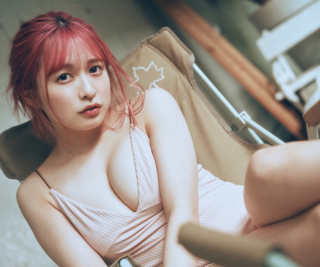 一名穿著淺色淺胸裙子的女性坐在椅子上，擁有粉紅色的頭髮，目光直視鏡頭，背景模糊且柔和。