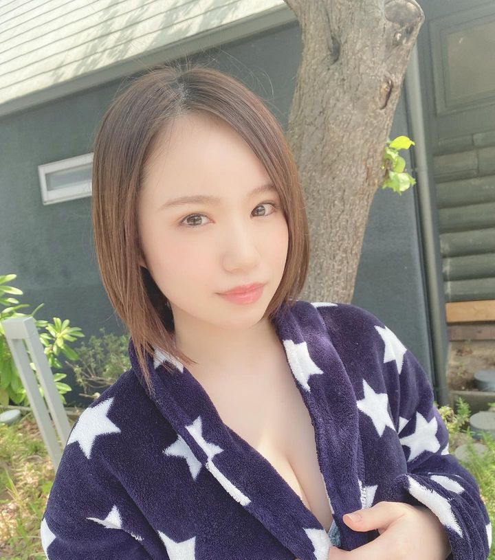 A young woman with short, straight hair wearing a dark robe with white stars, standing outdoors in front of a tree and a building.