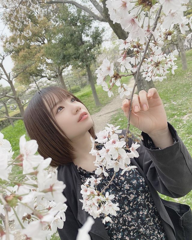 A young woman in a black jacket admires cherry blossom flowers while standing in a green park.