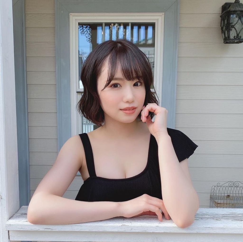 A young woman with wavy hair styled in a casual black top, resting her chin on her hand while leaning on a white railing in front of a house.