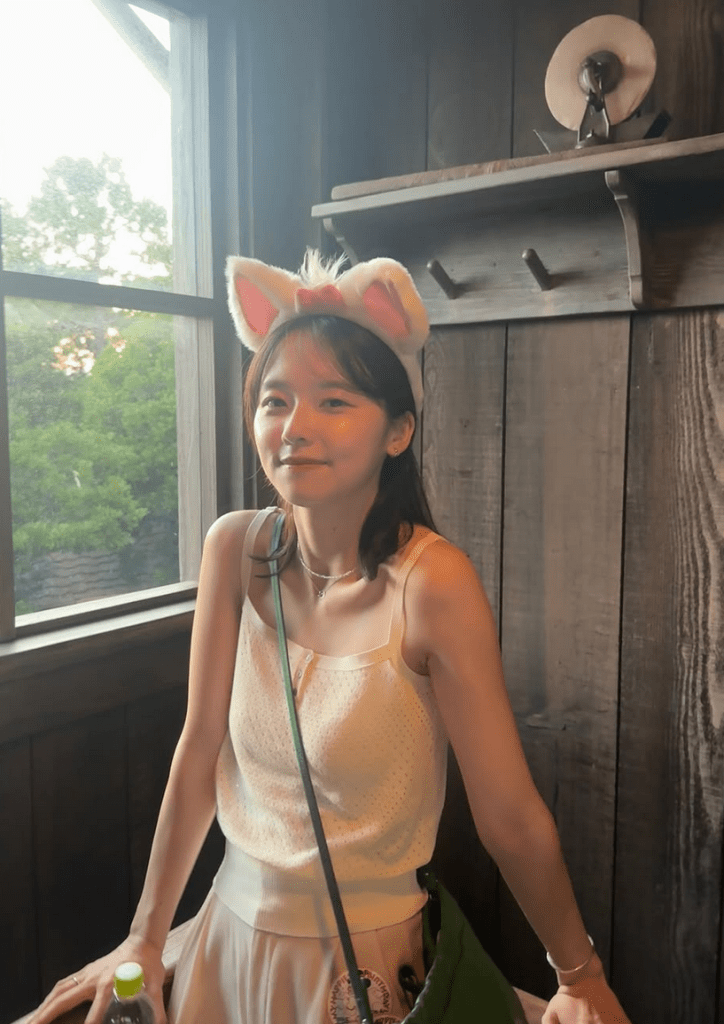 A young woman wearing a white tank top and a skirt, standing against a wooden wall with windows in the background. She has bunny ear headbands on her head and is holding a bottle of water.