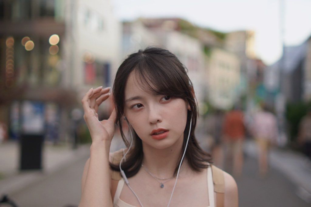 一位年輕女性在城市街道上，耳朵上插著耳機，神情思考，身後模糊的人群和建築物創造出動感背景。