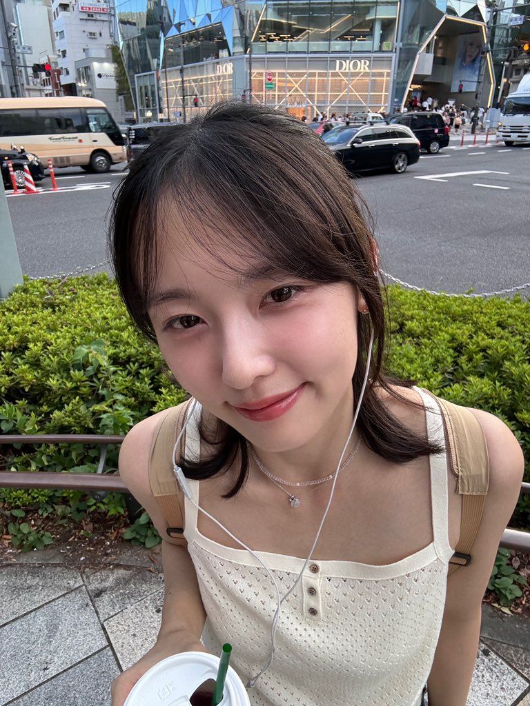 A young woman with long hair wearing a white top and earphones stands outdoors, smiling at the camera. In the background, a busy street and a high-end store can be seen.