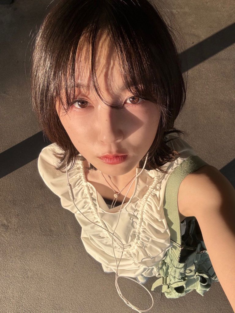 A young woman with short dark hair and a white ruffled top takes a selfie in sunlight, wearing earbuds and holding a green bag.