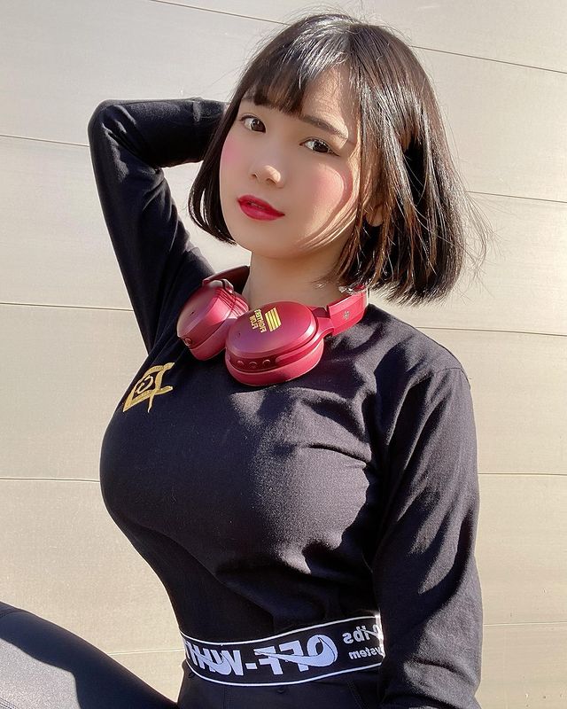 Young woman with short black hair wearing a black long-sleeve shirt and red headphones, posing with one hand on her head and the other at her side.