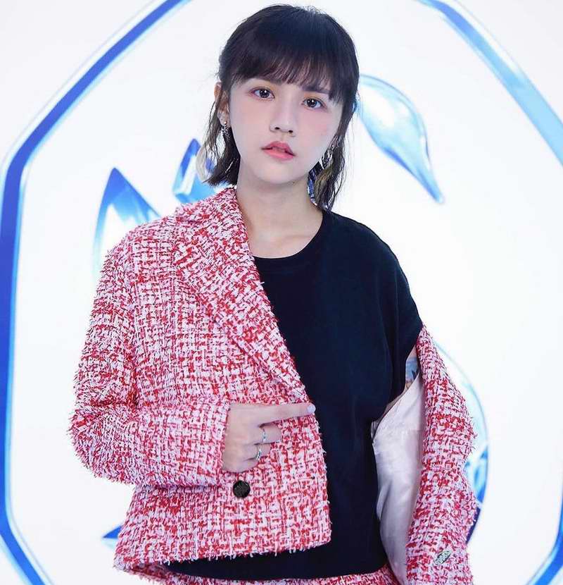 A young woman with dark hair and bangs is wearing a stylish red and white tweed blazer over a black top, posing in front of a blue abstract background.