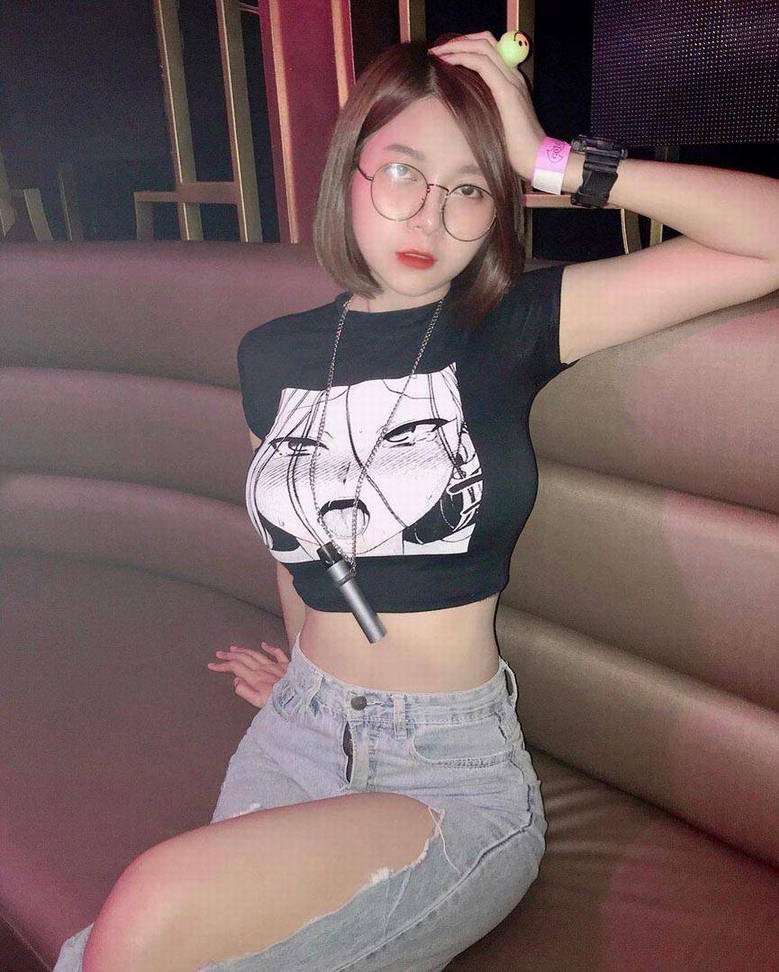 A young woman with short brown hair and glasses sitting on a couch, wearing a black crop top with an artistic print and light-wash ripped jeans.