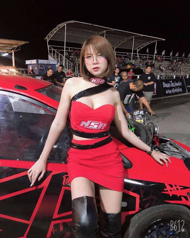 A woman in a red dress with a black strap posing next to a red race car, wearing glasses, with spectators in the background at a nighttime racing event.