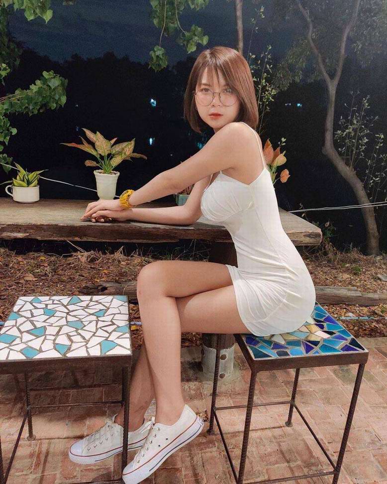 A young woman with glasses wearing a white dress sits on a stool, leaning forward with a serene expression. She is surrounded by foliage and a rustic wooden table with two mosaic-top stools.