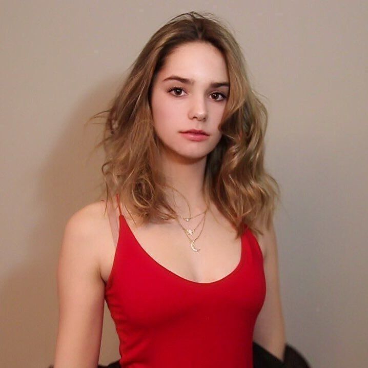 Portrait of a young woman with wavy hair wearing a red tank top, standing against a neutral background.
