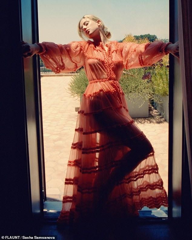 A woman in a flowing red dress stands at an open door, posing gracefully with her arms extended. The background features greenery and a sunny outdoor setting.