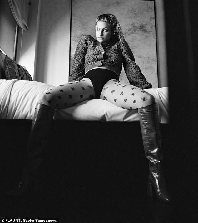 A woman sits on a bed in a stylishly posed manner, wearing a cropped sweater and patterned tights, with silver high-heeled boots and loose hair, captured in black and white.