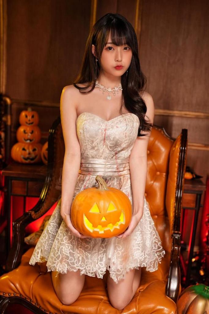 A young woman in a lace dress sits on a chair holding a carved pumpkin with a smiling face, surrounded by Halloween decorations.