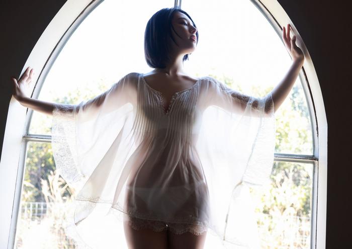 A woman standing in a window wearing a sheer, flowing garment, with her arms outstretched and a serene expression.