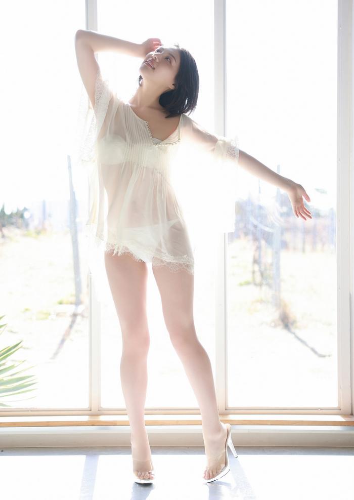 A woman in a sheer, light-colored outfit stands gracefully by a large window, with sunlight streaming in, creating a soft, ethereal effect.