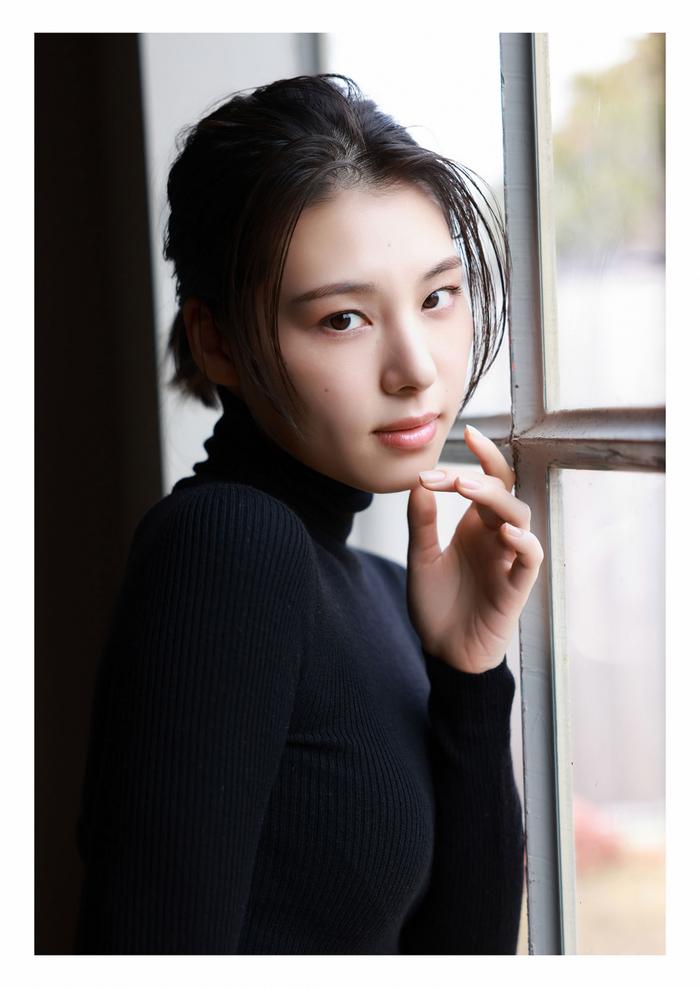 A young woman with long black hair styled in a casual updo is posing next to a window. She is wearing a black ribbed turtleneck sweater and has a thoughtful expression while gently touching her chin with one hand.