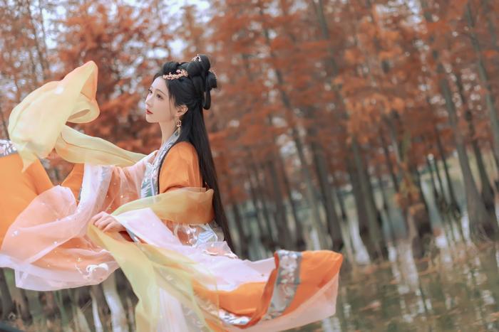 A woman wearing an orange and white traditional dress with flowing sleeves poses gracefully in an autumn landscape with vibrant orange trees.