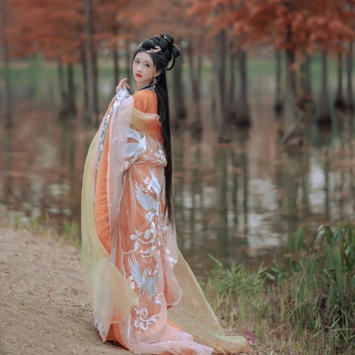 A woman in a traditional Chinese dress stands by a serene body of water, surrounded by autumn trees with orange leaves. The dress features intricate patterns and layers, enhancing her elegant pose and long black hair.