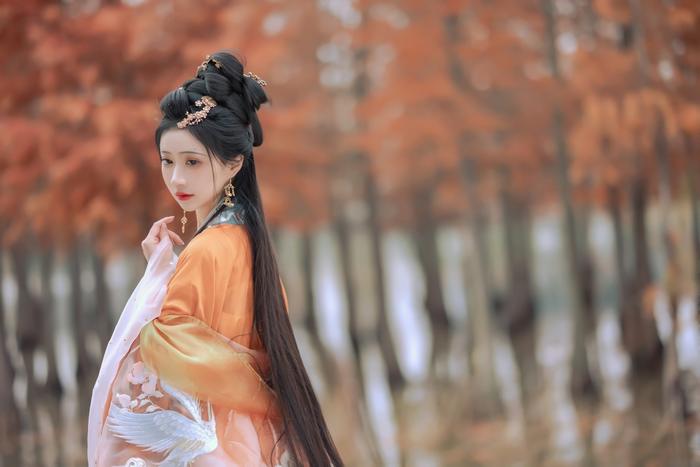 A young woman wearing an elegant orange traditional outfit stands in a forest with autumn leaves, displaying long hair and decorative accessories.